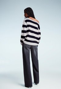 Braidelt hajú nő fekete-fehér csíkos, vállnélküli pulóverben és széles szárú sötét farmert visel, egy sima háttér előtt áll.