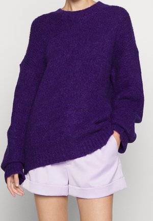Personne portant un pull tricoté épais violet à manches longues associé à un short lilas à ourlets retroussés, sur fond uni.