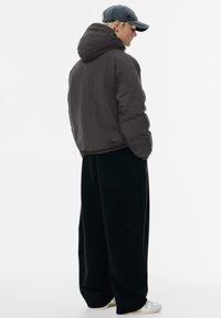 Donkergrijze pufferjas met een hoge kraag en capuchon; loszittende zwarte broek; sneakers met een lichte zool; gezien vanuit de achterkant.