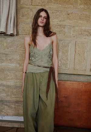 Femme aux longs cheveux bruns portant un caraco en dentelle verte et un pantalon à jambes larges, debout contre un mur en pierre texturée à l'intérieur.