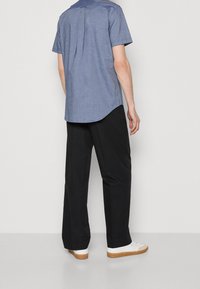 Homme debout de dos portant une chemise bleue à manches courtes, un pantalon noir et des baskets blanches avec semelles marron sur un fond uni.