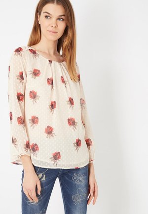 Joven mujer con una blusa blanca translúcida con estampado floral rojo y vaqueros azules bordados con abejas, de pie frente a un fondo claro y sencillo.