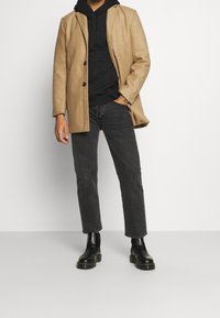 Manteau en laine beige à boutons noirs, porté sur un sweat à capuche noir. Jean slim noir et bottines noires complètent la tenue.