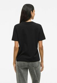 Svart bomull t-shirt med kort ärm och avslappnad passform. Bakifrån syns en rundad nertill och mjuk textur, tillsammans med byxor i grå vertikala ränder.