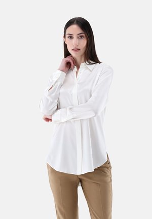 Camicia bianca a maniche lunghe con colletto button-down, tessuto liscio e orlo dritto, indossata con pantaloni beige. Bottoni visibili sulle maniche.