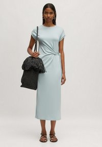 Lichtblauwe jurk tot de knie met korte mouwen en een knoopdetail bij de taille. Model houdt een zwarte, franjerugzak van geweven materiaal vast.