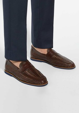 Brown leather loafers with woven design and smooth accents. Features a blue rubber sole and are paired with navy trousers.