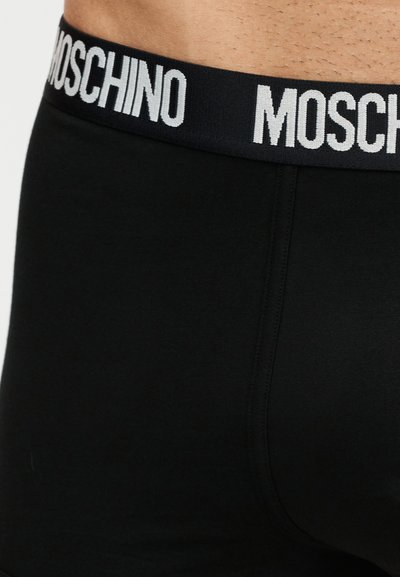 Κοντινό πλάνο μαύρων ανδρικών μπόξερ με μαύρη μέση και λευκό κείμενο «MOSCHINO», φορεμένα στη μέση ενός ατόμου.