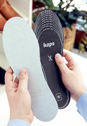 Hænder holder et par perforerede skosåler, den ene ensfarvet lys grå og den anden sort med størrelsesmarkeringer og en skærevejledning.