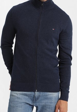 Homme portant un pull bleu marine zippé avec un petit logo sur la poitrine et un jean bleu clair, debout avec une main dans la poche.