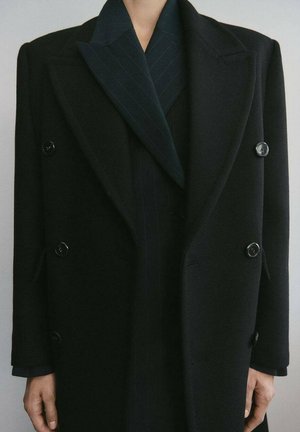 Osoba ubrana w czarny dwurzędowy wełniany płaszcz na ciemnym marynarce w cienkie prążki, ręce swobodnie opuszczone wzdłuż ciała na jednolitym tle.