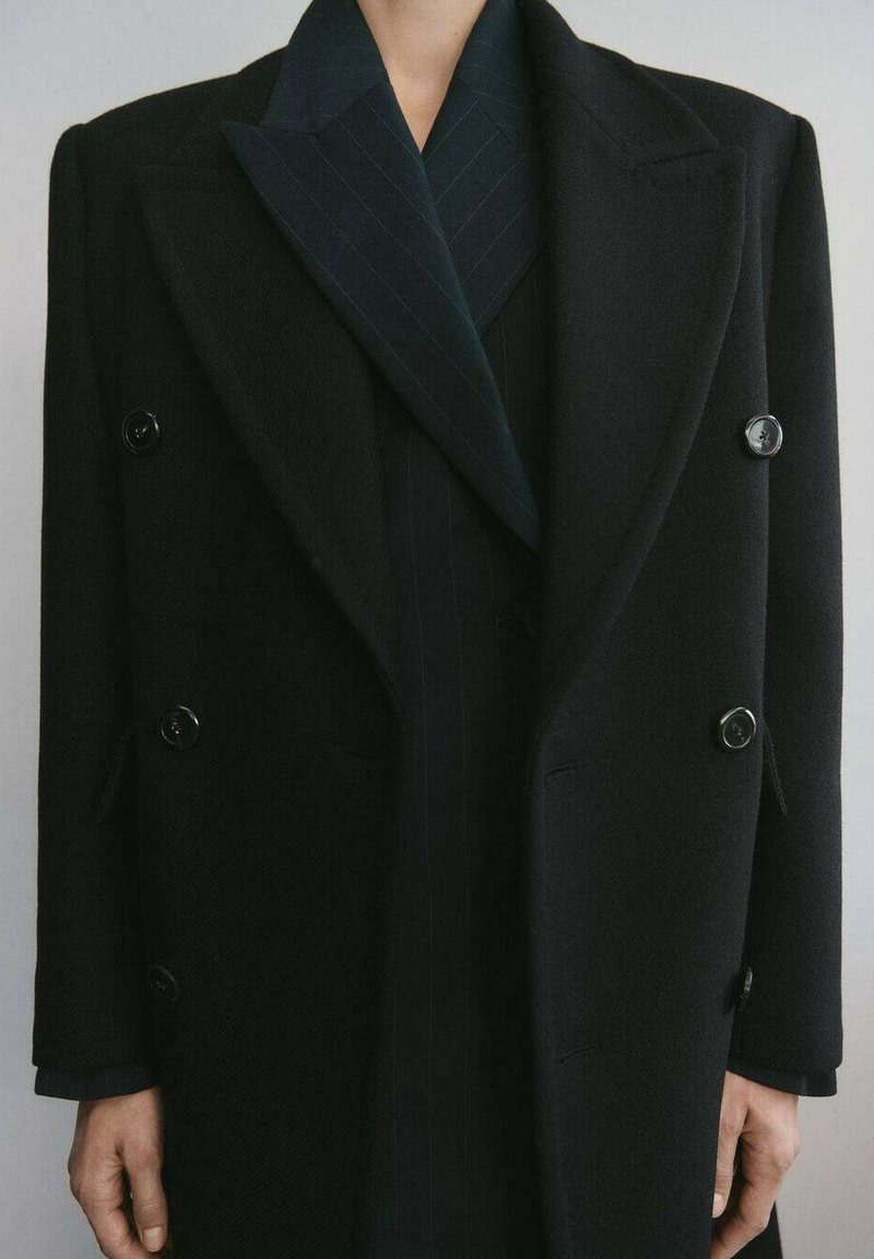 Persona vistiendo un abrigo negro de lana de doble botonadura sobre un blazer oscuro con rayas finas, con las manos relajadas a los lados sobre un fondo simple.