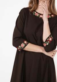 Kvinna i en mörkbrun klänning med röd, krämfärgad och grön blommig broderi vid halsringning och ärmarnas manschetter, poserar med armarna i kors.