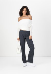 White off-the-shoulder top, long sleeves, paired with dark pinstriped trousers and white sneakers. Model holds a small white handbag.