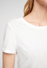 Gros plan d'une personne portant un t-shirt blanc uni à manches courtes avec un col rond et des manches retroussées.