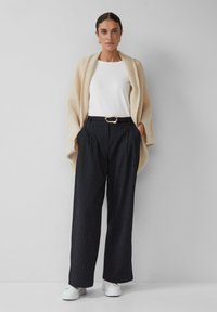Châle texturé crème sur une chemise blanche, associé à un pantalon noir à rayures verticales taille haute et des baskets blanches. Détail de boucle de ceinture en or visible.