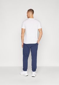 Balti trumpai rankovėmis marškiniai, derinami su tamsiai mėlynomis elastinėmis kelnių sportinėmis kelnėmis. Modelis vilki baltus sportinius batelius. Švarus, minimalus dizainas, be matomų raštų.