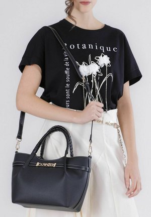 Donna con t-shirt nera con motivo floreale e gonna bianca, che porta una borsa nera in pelle con manici intrecciati e cintura con catena dorata.