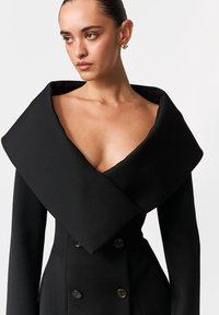 Czarny dwurzędowy płaszcz z szerokim, podwiniętym kołnierzem. Wykonany z teksturowanej tkaniny, z metalicznymi guzikami i usztywnionymi rękawami.