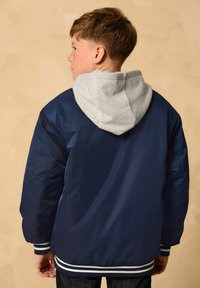 Dunkelblaue Jacke mit grauem Kapuzen, elastischen Bündchen und gestreiftem Saum. Glatte Textur mit genähten Details. Getragen von einem Kind, von hinten betrachtet.