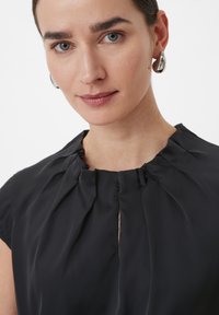 Blouse noire à col montant, avec un tissu froncé et un détail d'ouverture sur le devant. La texture lisse et les manches courtes complètent le design.