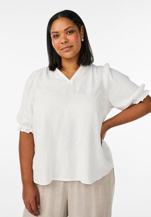 Woman with shoulder-length black hair wearing a white V-neck blouse with puff sleeves and beige pants, posing with one hand on her hip.