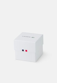 Baltas kvadratinis dėžutė su vyriška dangčiu, kuriame yra LACOSTE logotipas. Priekyje puošia mėlynos, raudonos ir baltos juostos. Lygus paviršiaus tekstūra.