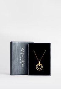 Guldkedja med två sammanflätade ringar, en av dem inlagd med en liten rund diamant, presenterad i en svartsammetfodrad låda. Silverlogotyp på lådan.