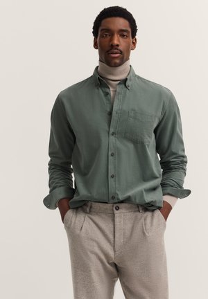 Mann mit grünem Hemd über beige Rollkragenpullover und hellgrauen Hosen, steht mit Händen in den Taschen vor einfarbigem Hintergrund.