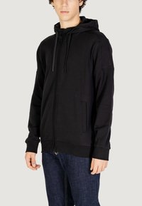 Felpa nera con zip, dal texture morbido, con cappuccio regolabile e tasche laterali. Presenta polsini e orlo a coste, progettata per un fit rilassato.