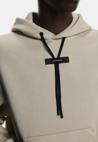 Hoodie beige avec une texture douce, présentant un cordon noir et une étiquette portant la mention "INGÉNIERIE SUISSE." Poche kangourou avant visible.