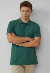 Koszulka polo w ciemnozielonym kolorze, wykonana z bawełny, z krótkimi rękawami, zapięciem na trzy guziki, prostym dołem i luźnym krojem. Prosty design, bez wzorów ani logo.