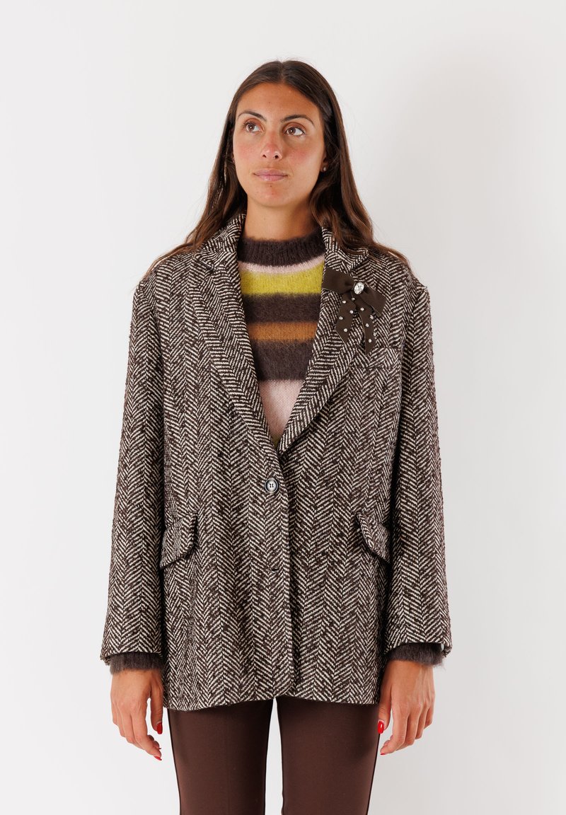 Blazer marrone a motivo houndstooth con due tasche, revers a punta e bottoni, indossato sopra un maglione a righe marrone e giallo.