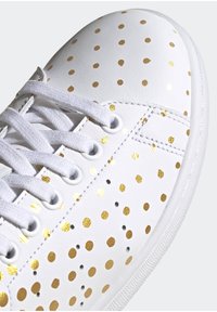 Vita lädersneakers med ett prickigt mönster i guld och svart, med vita snören och öglor, samt en texturerad gummisula.