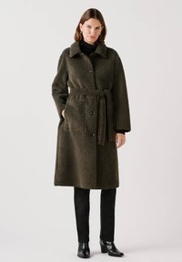 Olijfgroene, geëxtraheerde lange jas met een kraaglijn, knoopsluiting, ceintuur in de taille en twee voorzakken. Gepaard met zwarte broek en schoenen.