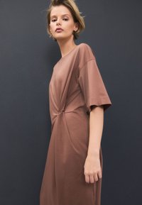 Brun klänning med korta ärmar, mjukt tyg, avslappnad passform med en tvistdetalj vid midjan, slät textur, bärs mot en mörk bakgrund.