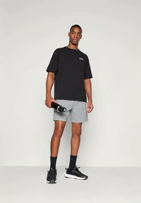 Μαύρη κοντομάνικη μπλούζα και γκρι σορτς, κρατώντας μία μαύρη μπουκαλιέρα νερού. Μαύρα αθλητικά παπούτσια με υφή στη σόλα. Μινιμαλιστικός σχεδιασμός.