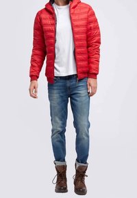 Röd puffjacka med horisontell quiltad design, dragkedja och huva; bärs över en vit T-shirt och kombineras med blå jeans och bruna kängor.