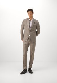 Costume beige clair avec un design ajusté, fermeture à un bouton et revers en pointe. Associé à une chemise blanche et des chaussures de ville noires.