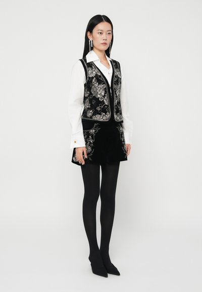 Femeie îmbrăcată cu o vestă neagră brodată cu flori și fustă, cămașă albă cu mâneci lungi, colanți negri și tocuri negre ascuțite, stând în fața unui fundal alb.