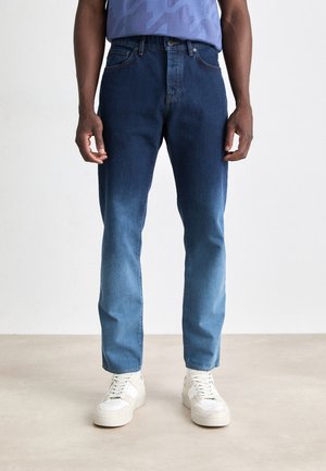 Mężczyzna w dżinsach w dwóch odcieniach niebieskiego i białych trampkach, stojący na jasnym, jednolitym tle.