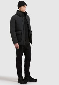 Schwarze gepolsterte Jacke mit hohem Kragen, Reißverschluss vorne und Seitentaschen; getragen über einem schwarzen Pullover und enganliegenden Hosen mit schwarzen Schnürstiefeln.