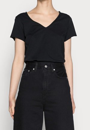 Frau trägt ein schwarzes, kurzärmeliges Shirt mit V-Ausschnitt, das in hoch taillierte schwarze Jeans mit Fronttaschen und silbernen Knöpfen gesteckt ist.