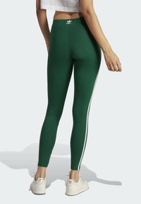 Gröna leggings i stretchmaterial med hög midja och vita sidostrimlor. Modellen har på sig vita sneakers med en logotyp.