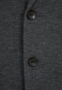 Camisa gris de tela con botones, acabado texturizado, que presenta dos botones redondos negros y un diseño limpio, enfatizando la simplicidad y funcionalidad.