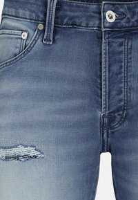 Gros plan sur un jean en denim bleu avec une petite déchirure usée près de la poche gauche et des détails de couture visibles.