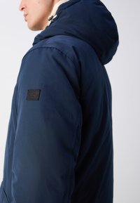 Chaqueta azul marino con capucha, que presenta una tela texturizada y una etiqueta negra en la manga. El diseño es funcional y aerodinámico.