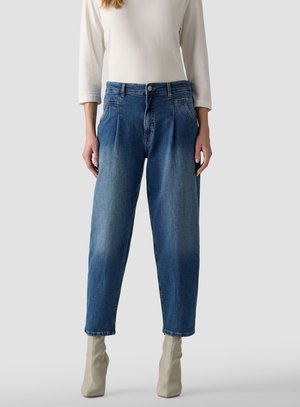 Woman wearing high-waisted, tapered blue jeans with pleats paired with beige ankle boots and a white long-sleeve top against a plain background.