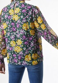 Blusa floreale in tessuto verde trasparente con rose gialle e viola. Maniche lunghe a sbuffo con polsini elastici e dettaglio a occhiello sulla schiena.