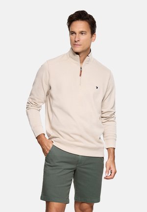 Hombre con sudadera beige con cremallera hasta el pecho y pantalones cortos verde oliva, de pie con una mano en el bolsillo contra un fondo liso.